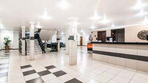 Um hall amplo com piso xadrez, balcão de recepção, escada e um relógio grande na parede.