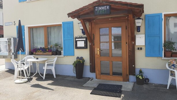 Un edificio con una puerta de madera que dice "Zimmer Frei" y un pequeño espacio al aire libre con sillas blancas y una mesa.