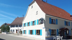 Un edificio de dos pisos con contraventanas azules, un letrero que indica 'WALDHORN', y un pequeño espacio al aire libre con sillas de plástico blanco.