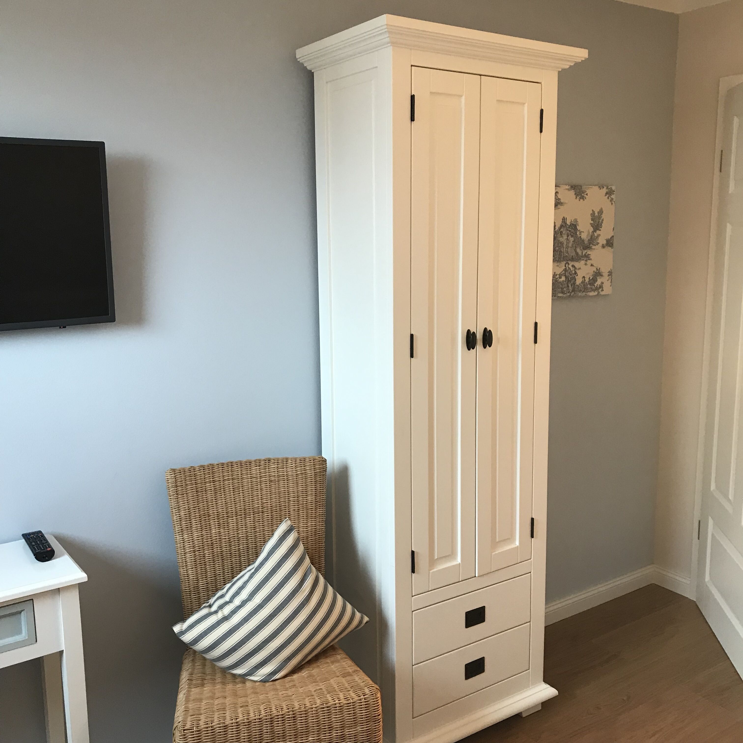 Ein Zimmer mit einem hohen, weißen Kleiderschrank, einem Korbstuhl mit gestreiftem Kissen, einem kleinen weißen Tisch mit Fernbedienung und einem gerahmten Bild an der Wand.