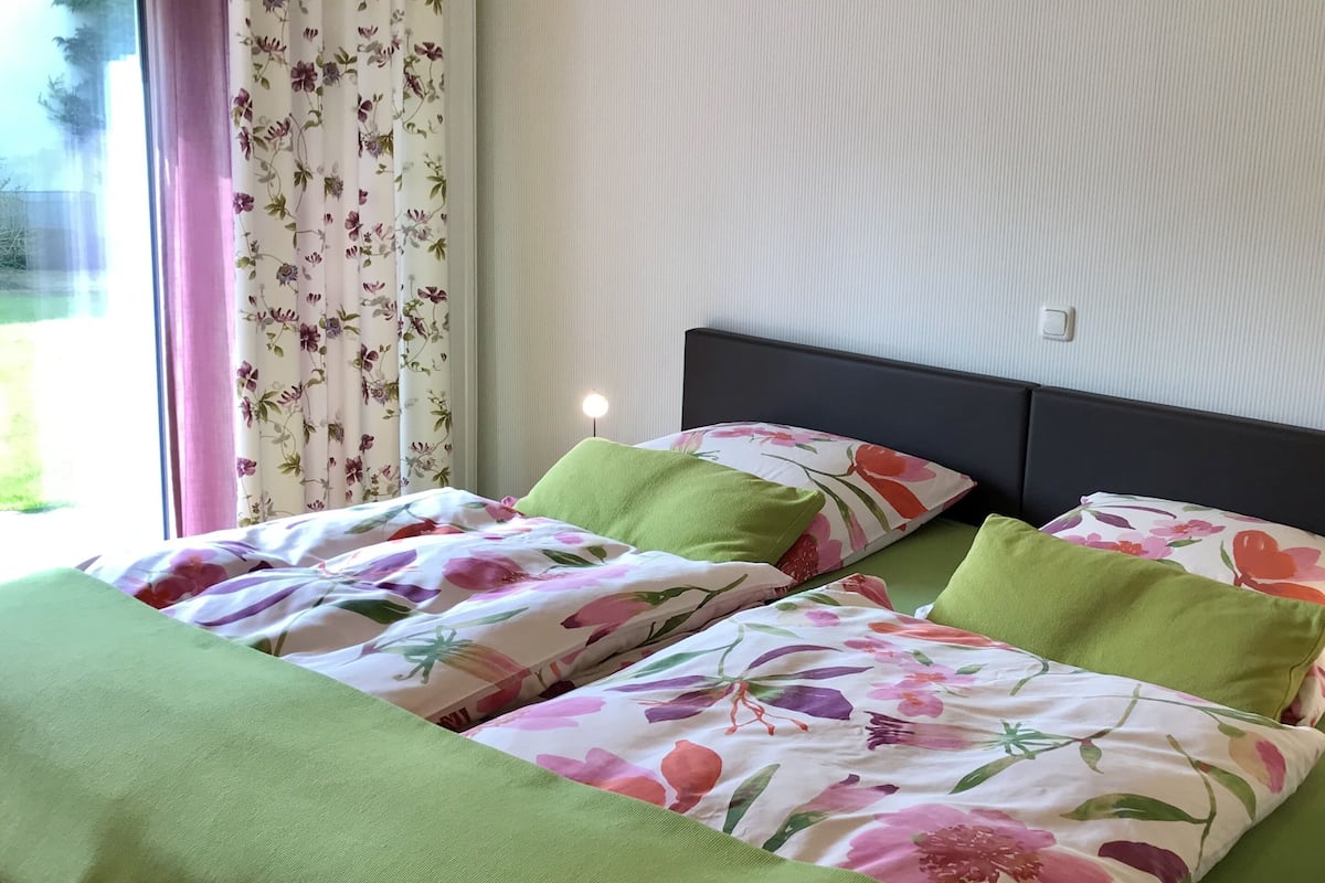 Cama doble con ropa de cama floral y almohadas verdes, mesita de noche con lámpara y ventana con cortinas florales.