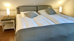A neatly made bed with striped bedding, a bedside table with a lamp, and a wooden nightstand. - Inselferien Micheel (Fehmarn)