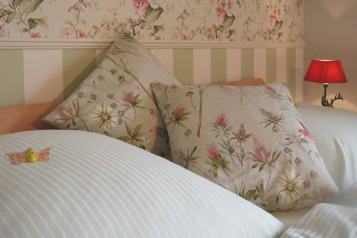 Un dormitorio con papel tapiz floral, cabecero a rayas y una lámpara roja sobre una mesita de noche.