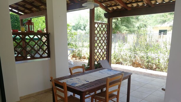 A covered patio area with a wooden table and chairs, a window with a view of greenery, and a tiled floor. - Villa Serena - Torre Inserraglio Nardò (Nardò)