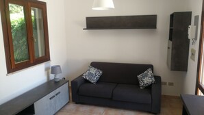 A modern living room with a dark grey sofa, two patterned cushions, a white pendant light, a built-in cabinet, and a window with a view of greenery. - Villa Serena - Torre Inserraglio Nardò (Nardò)