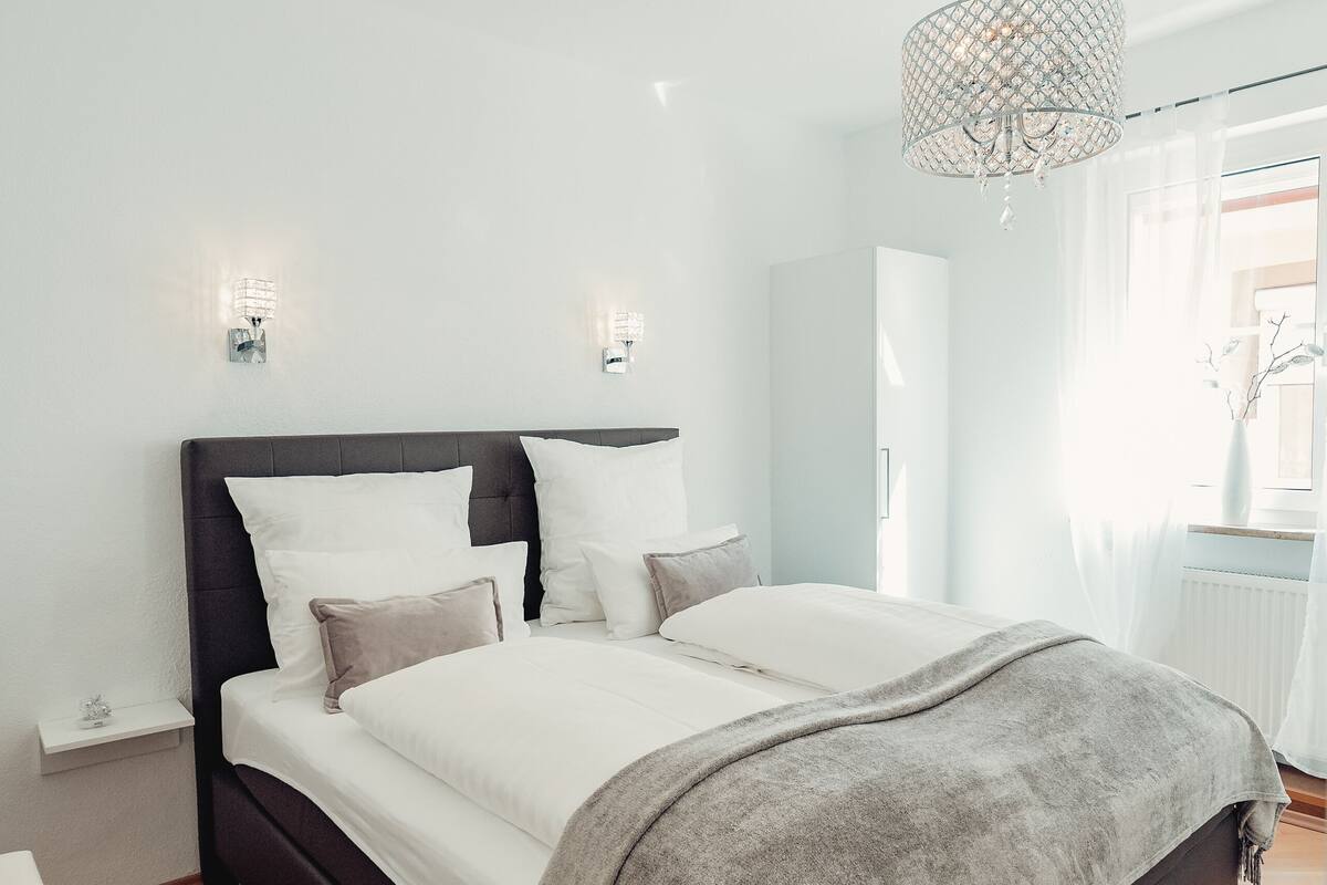 A modern bedroom with a large bed, a bedside table, a nightstand, and a hanging light fixture.