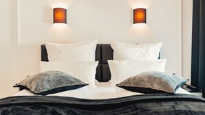 Un lit avec des oreillers blancs et gris, une tête de lit noire et deux lampes fixées au mur.