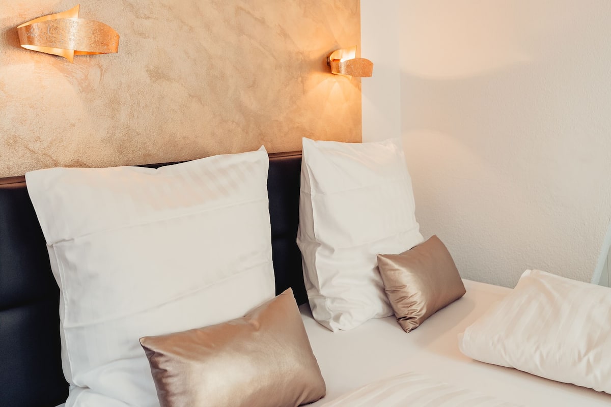 A hotel room with a textured ceiling, a dark headboard, and a bed with white and gold pillows.