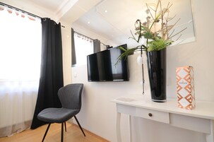 Una habitación moderna con televisión de pantalla plana, una silla negra, un escritorio blanco, una planta en maceta y una caja con la marca Ferrari.