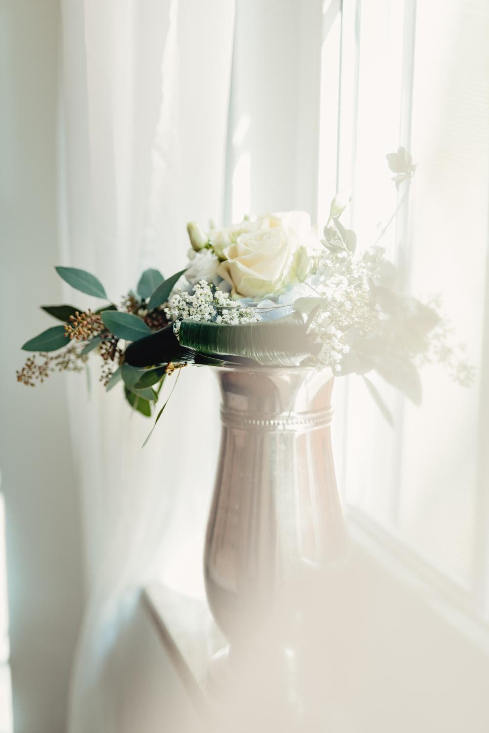 Un jarrón con un arreglo floral sobre un pedestal, cerca de una ventana con cortinas traslúcidas.