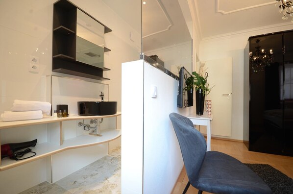 A modern bathroom with a glass partition, a white sink, and a black cabinet. There is a chair and a small table with a plant. - Hotel Da Felice (Bopfingen)