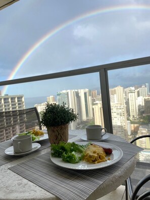 A dining table with a plate of food, a cup, and a potted plant on a balcony with a city view and a rainbow.