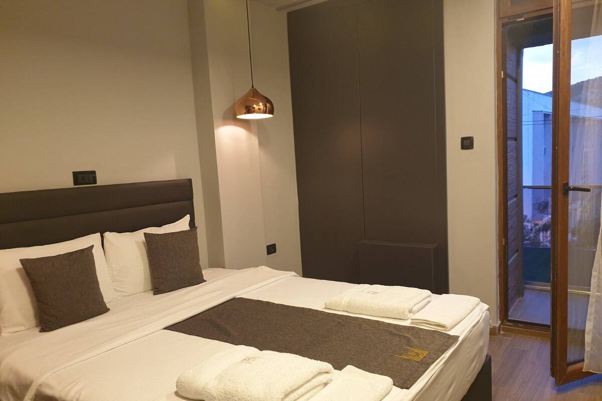 A modern hotel room with a large bed, a dark headboard, a hanging pendant light, and a view through a glass door.