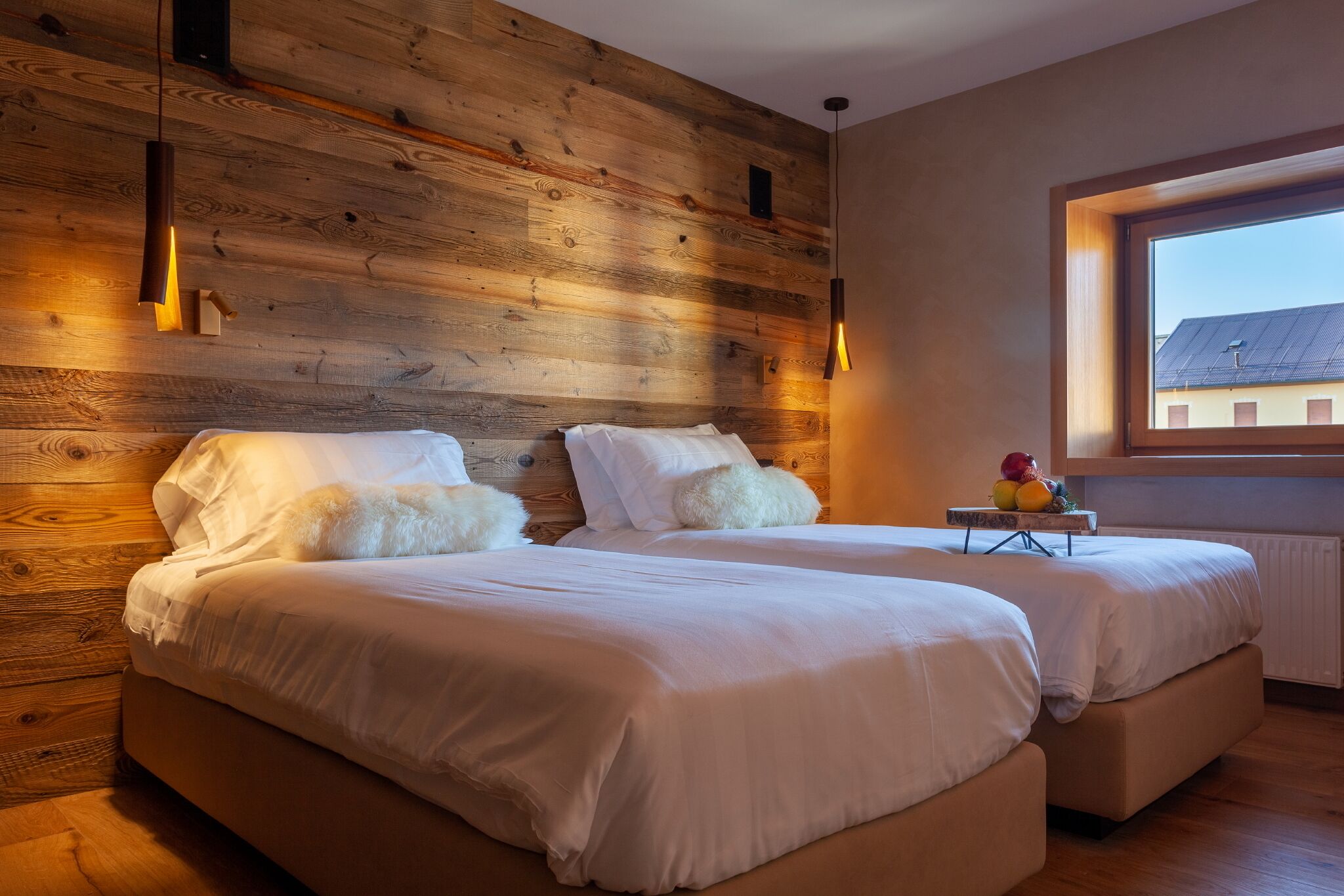 A bedroom with two beds, wooden wall paneling, pendant lights, and a window with a view of buildings.
