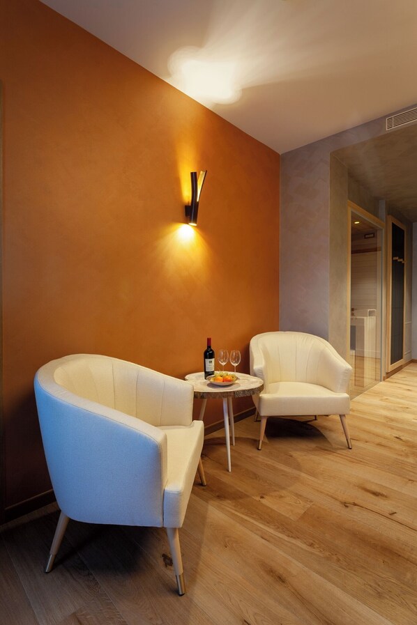 A modern room with two white armchairs, a small round table with a bottle and glasses, and a wall-mounted light fixture. - Asiago Sporting Hotel & SPA - Buongiorno! Hotels (Asiago)