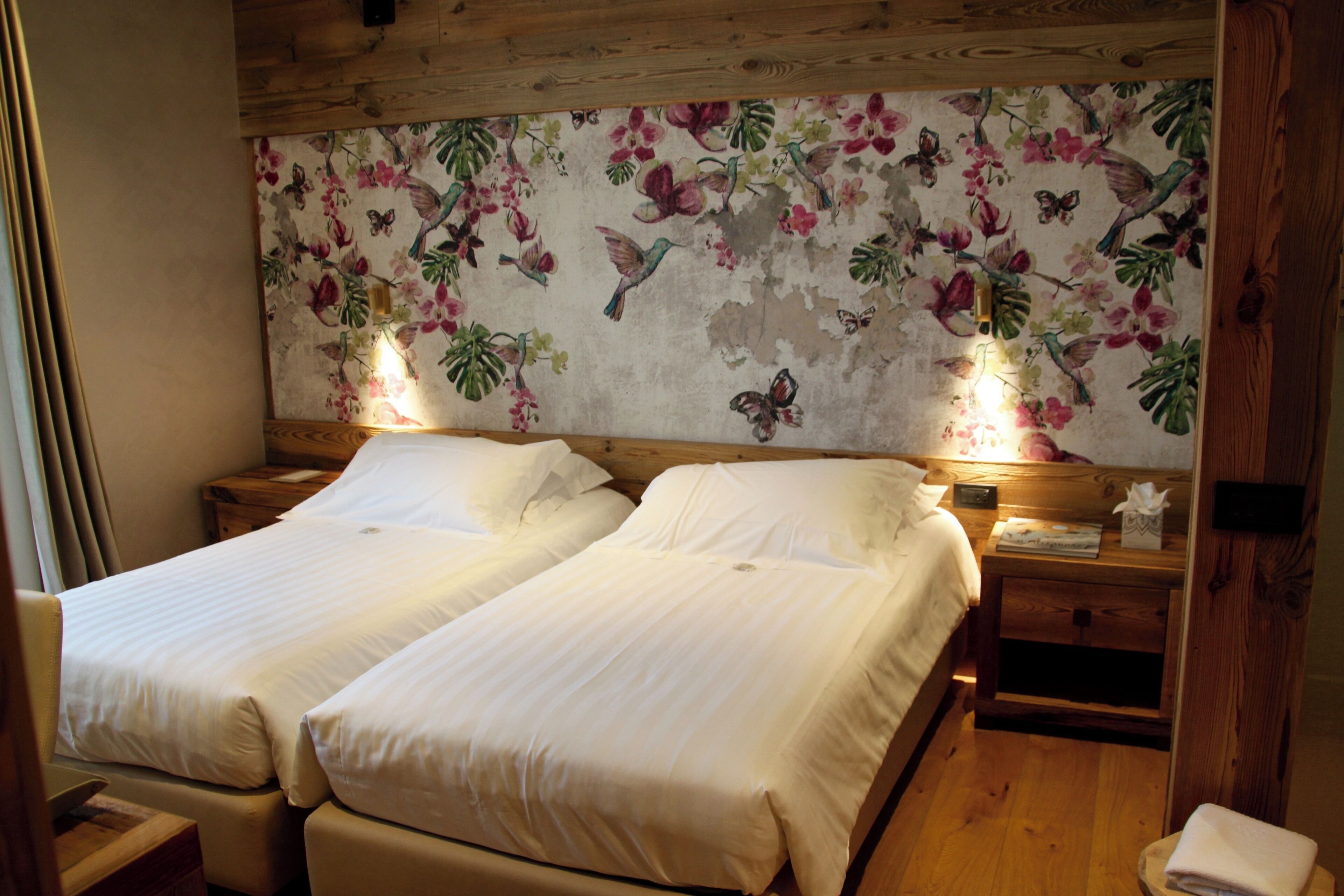 Una habitación con dos camas individuales, cabecera de madera, papel tapiz floral, lámparas de noche y una mesita de noche de madera con un dispenser de pañuelos.