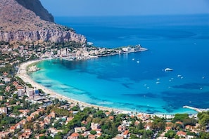 An aerial view of a coastal town with a beach, clear blue waters, and a rocky mountain in the background.