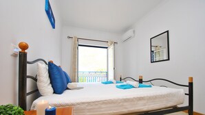 Una cama bien hecha con almohadas azules y blancas, una mesita de noche con una vela, y vistas a un balcón con barandilla.