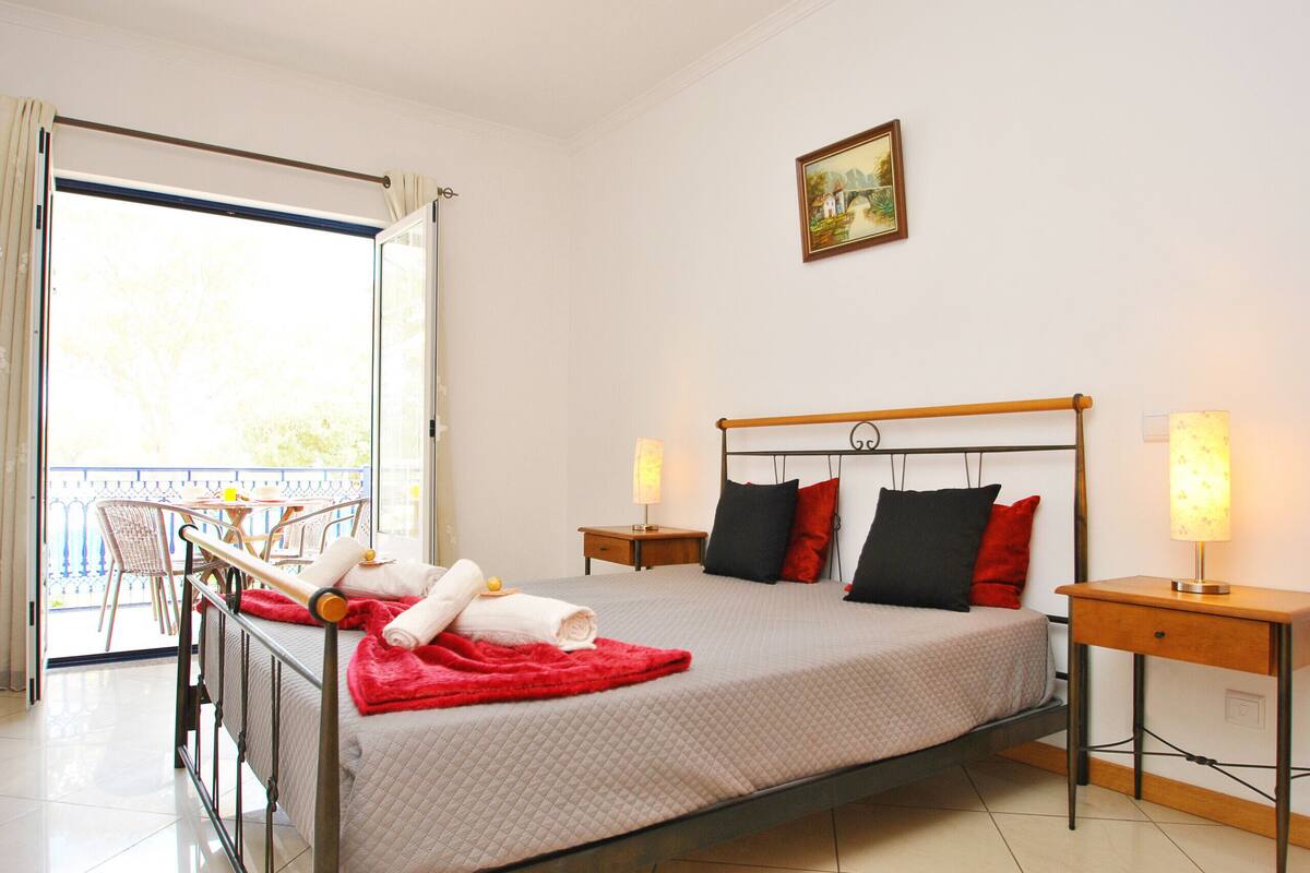 A neatly made bed with a red blanket and pillows, a bedside table with a lamp, and a balcony with chairs and a table.