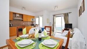 Un comedor con una mesa preparada para cuatro, una cocina al fondo y un balcón con televisor.