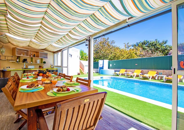 A dining area with a pool view, wooden table, chairs, and a striped awning.
