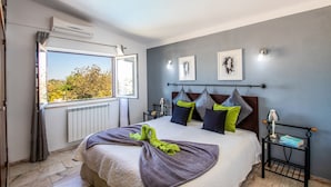 Un dormitorio con una cama grande, vistas a árboles a través de la ventana, un radiador y una mesita de noche con una lámpara.