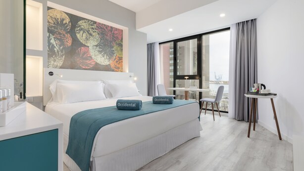 Habitación de hotel moderna con una cama grande, un escritorio y vistas a la ciudad a través de un amplio ventanal.
