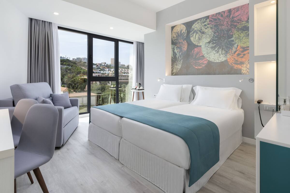 Una habitación de hotel moderna con una cama grande, un sofá, un escritorio y vistas de la ciudad a través de amplias ventanas.