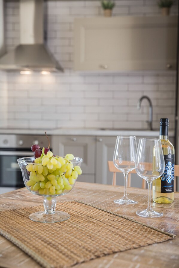 Una cocina con salpicadero de azulejos blancos, una campana extractora de acero inoxidable, una botella de vino blanco y un bol de uvas amarillas sobre un mantel tejido.