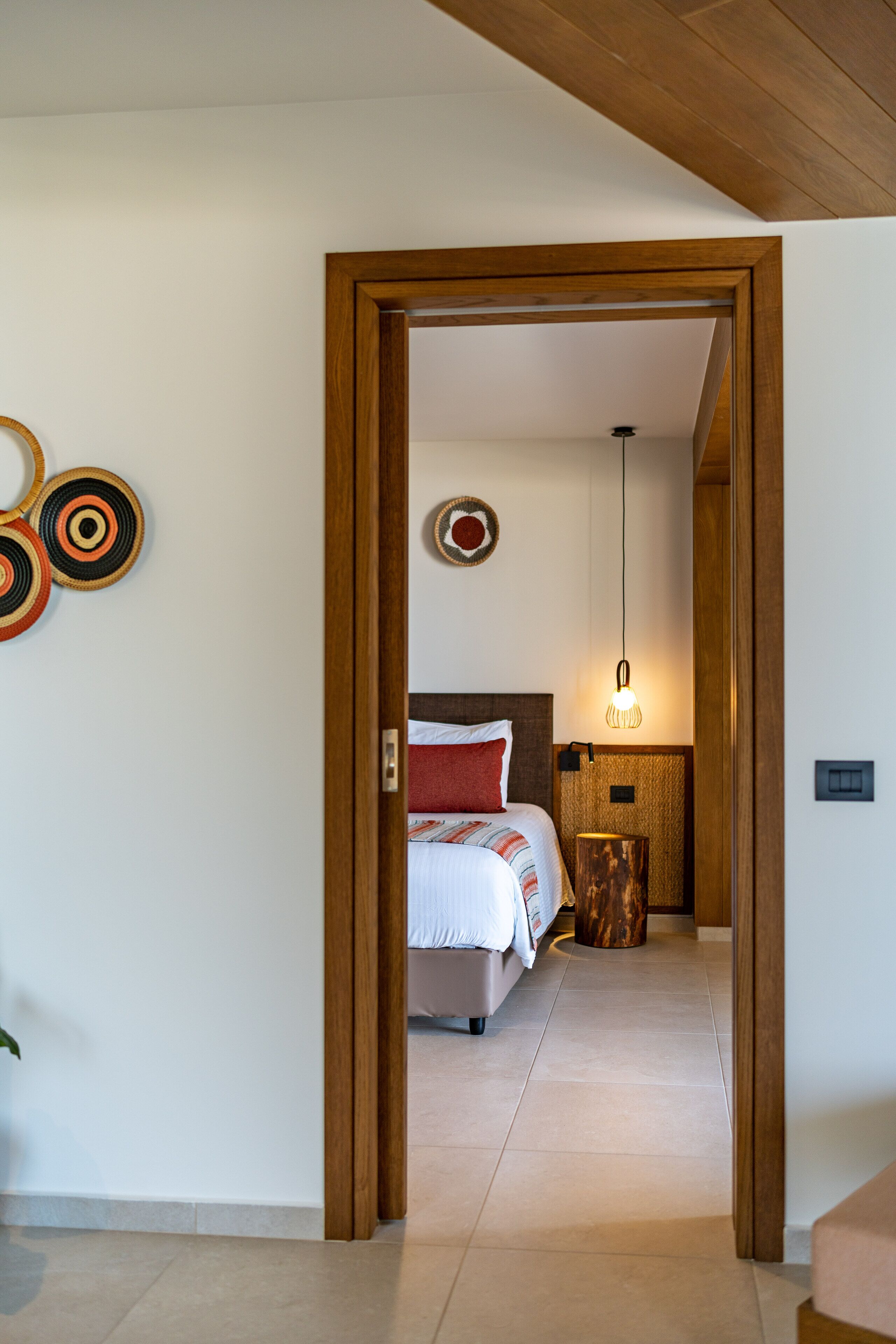 A bedroom with a bed, a bedside table, a wall-mounted light, and a door leading to another room.