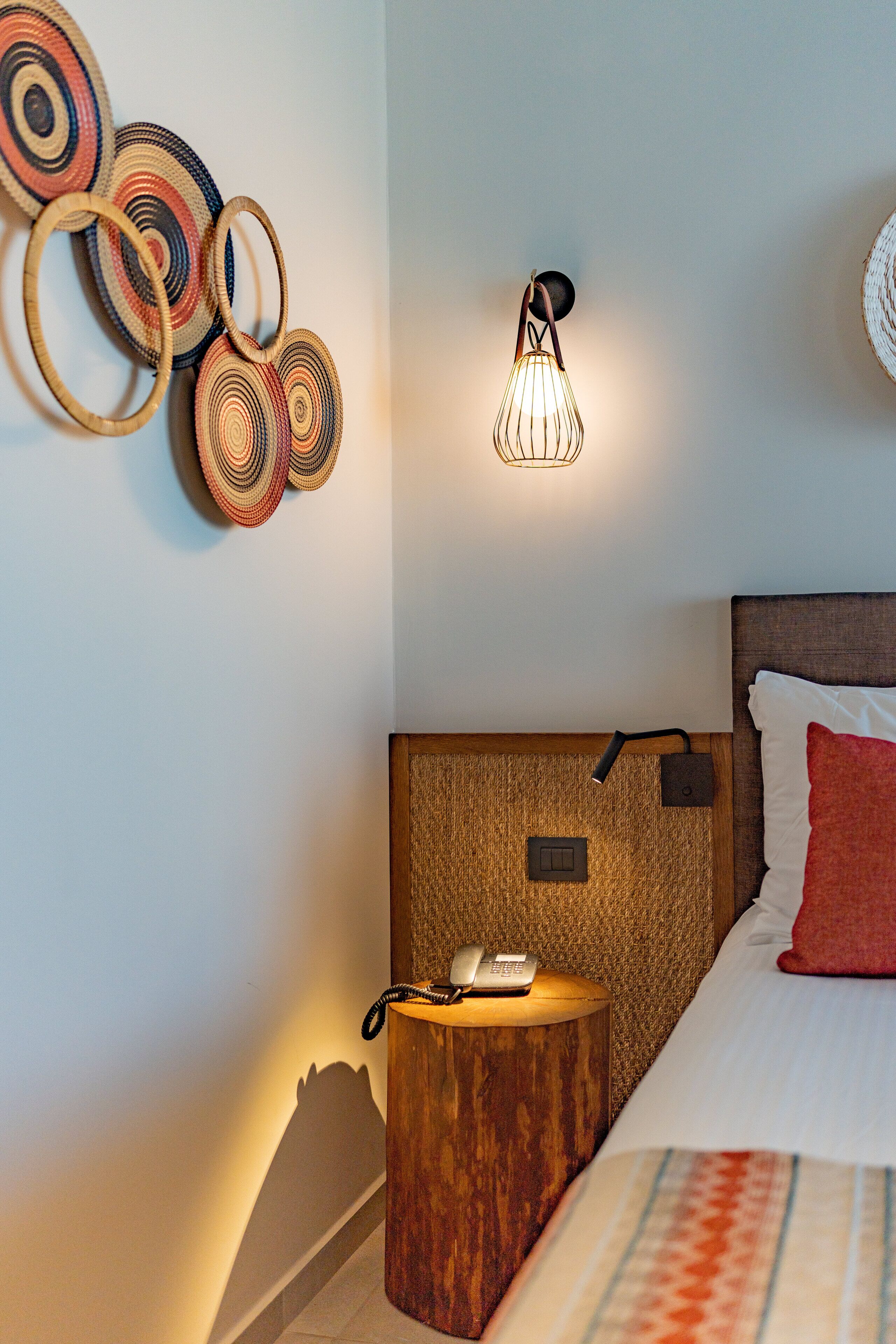 A hotel room with a wooden headboard, a bedside table with a phone, a wall-mounted light fixture, and a bed with a red pillow.