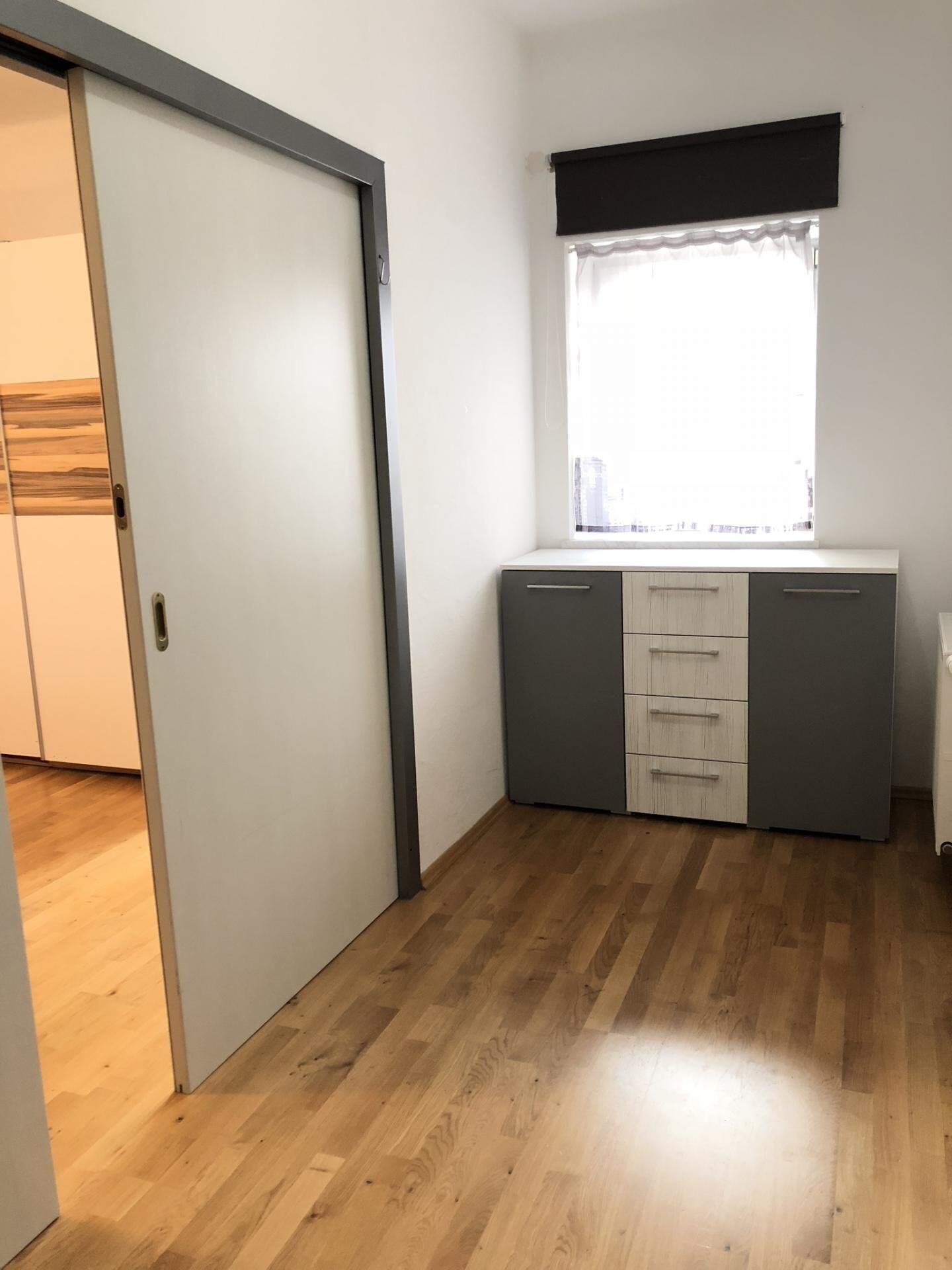 A small, bright room with a wooden floor, a sliding door, a window with a black valance, and a kitchenette area with cabinets and a countertop.