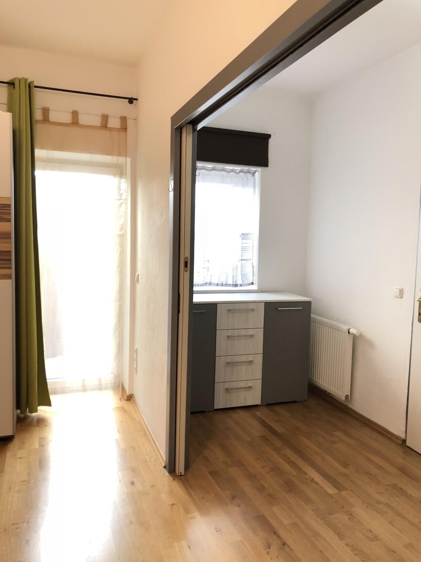 A room with a sliding door leading to a balcony, a window with a view of buildings, a radiator, and a small kitchenette area.
