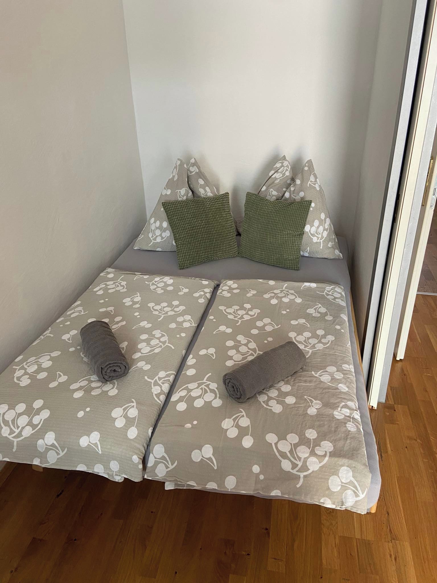 A small, single-bed room with a floral-patterned bedspread, two pillows, and two rolled towels.
