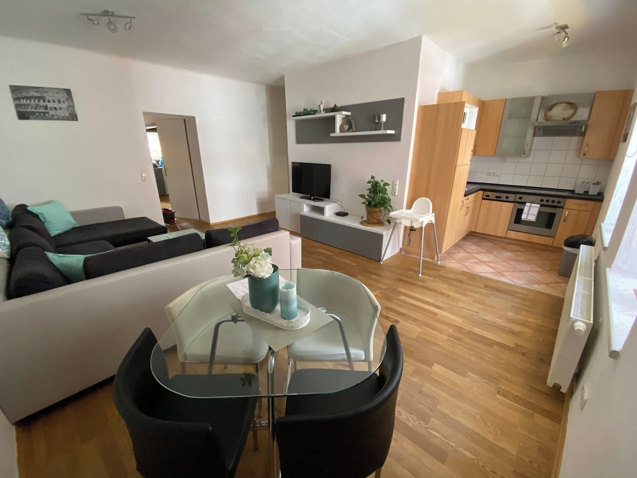 Uma sala de estar moderna com sofá, mesa de jantar com cadeiras, TV e área de cozinha com armários e fogão.