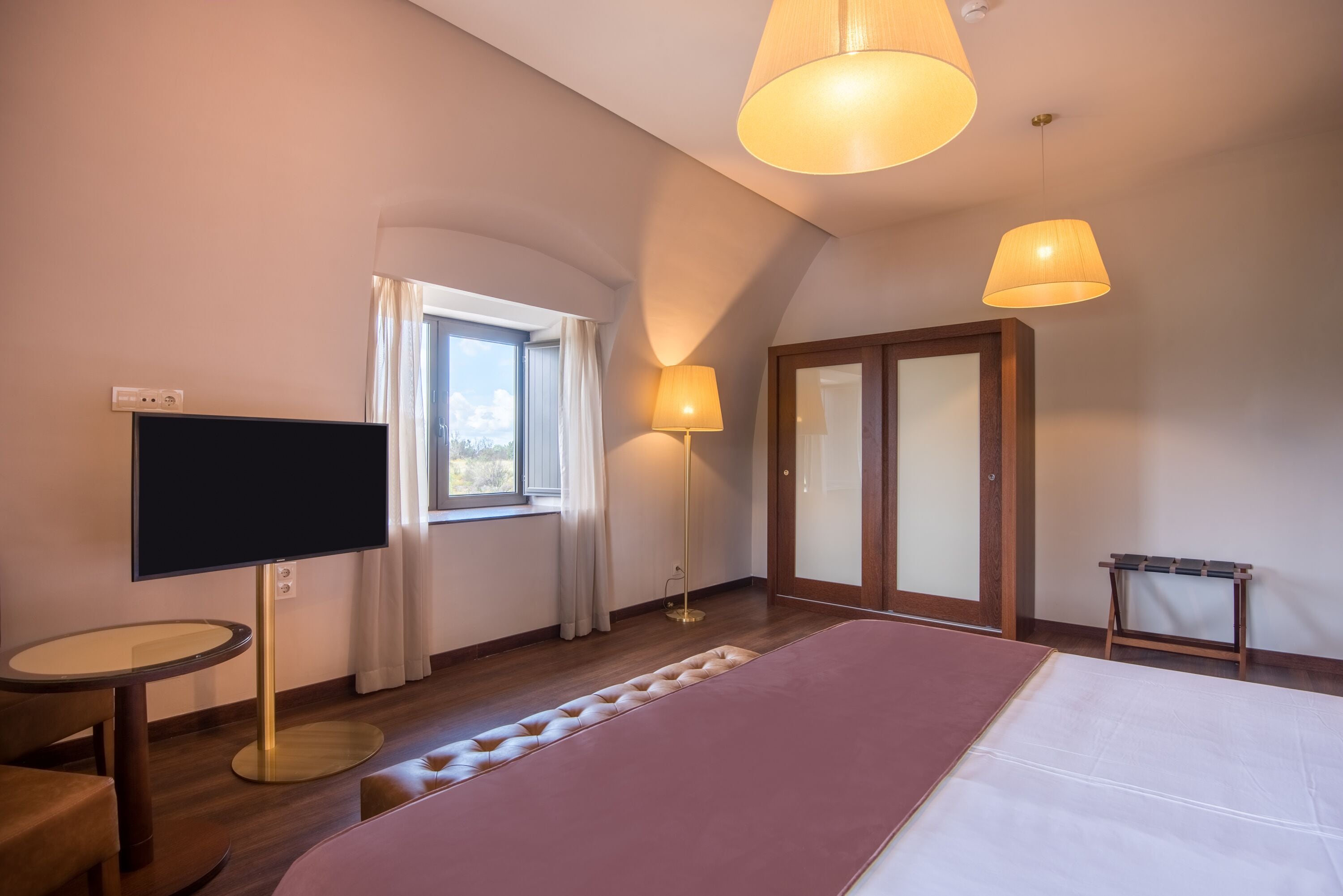 A modern hotel room with a large bed, a flat-screen TV, a round table, and a ceiling light.
