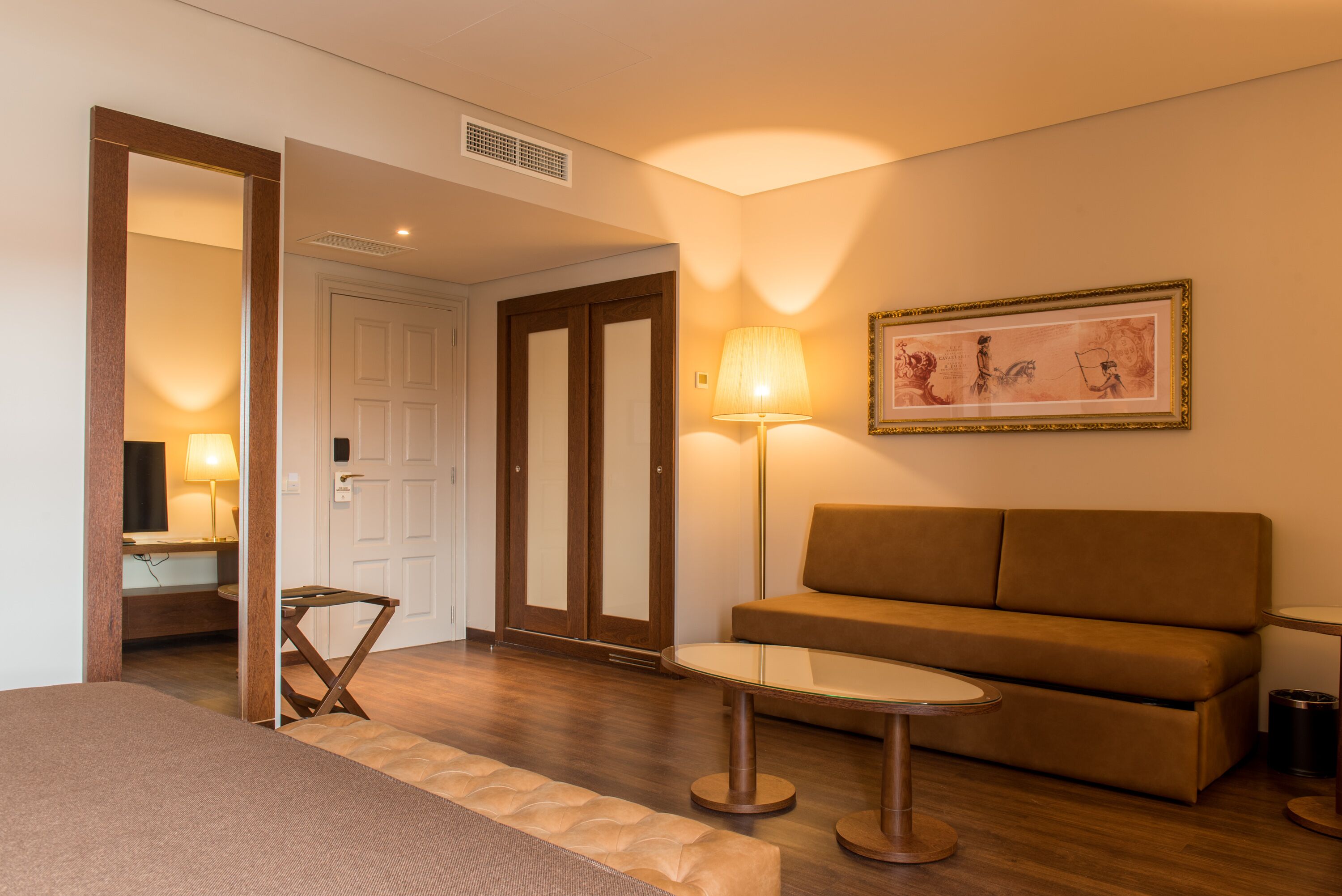 Habitación de hotel con suelo de madera, un sofá marrón, una mesita redonda pequeña, una lámpara y un cuadro en la pared.