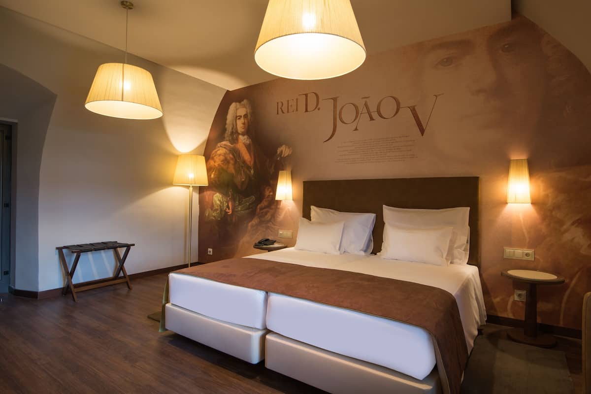 A modern hotel room with a large bed, two bedside lamps, a small table, and a wall-mounted artwork.