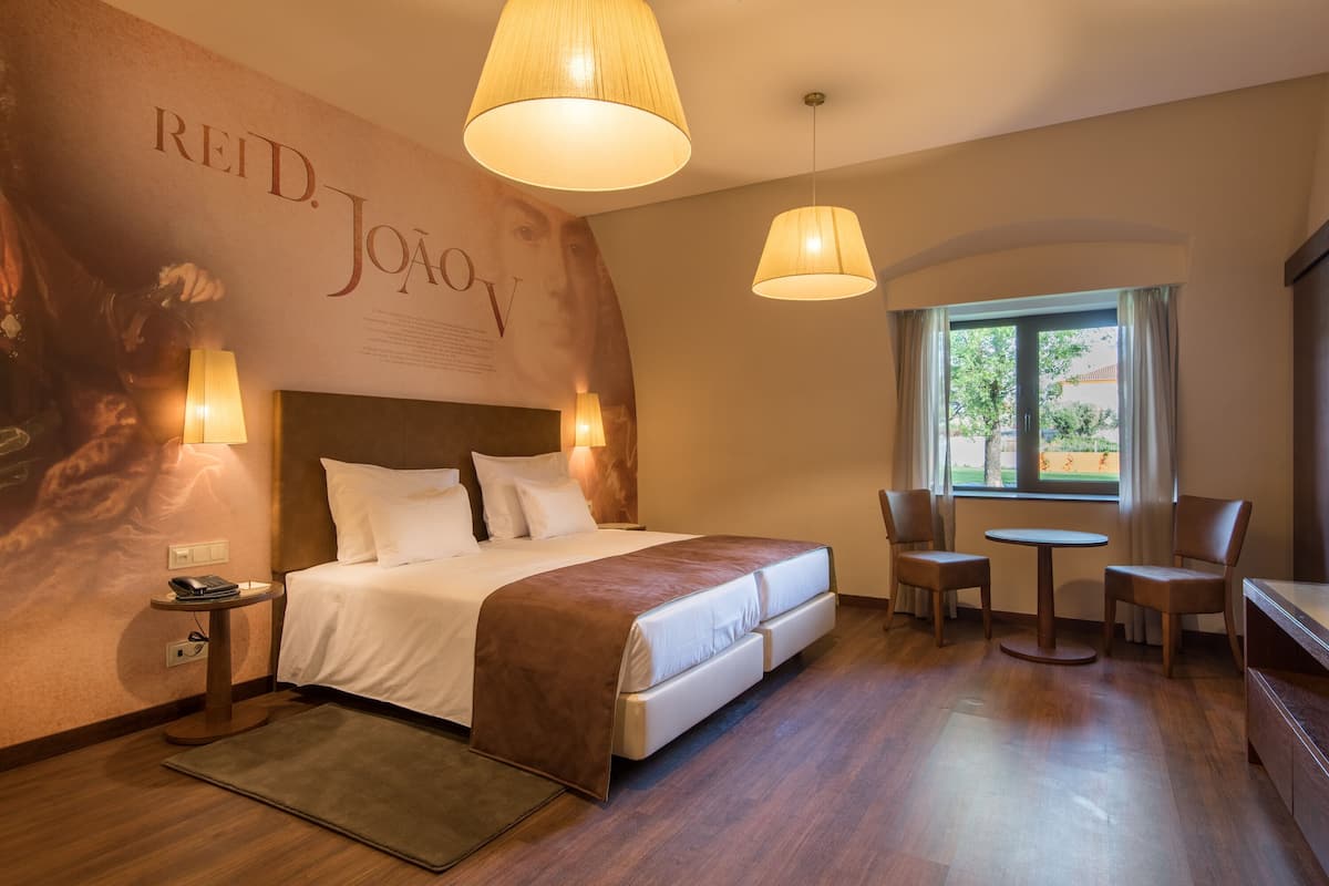 A hotel room with a large bed, two bedside lamps, a small table with chairs, and a window with a view of greenery.
