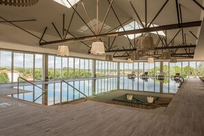 A large indoor swimming pool with clear views of the surrounding landscape, a wooden deck area, and modern pendant lighting. - Vila Gale Collection Alter Real - Resort Equestre, Conference & Spa (Alter do Chao)