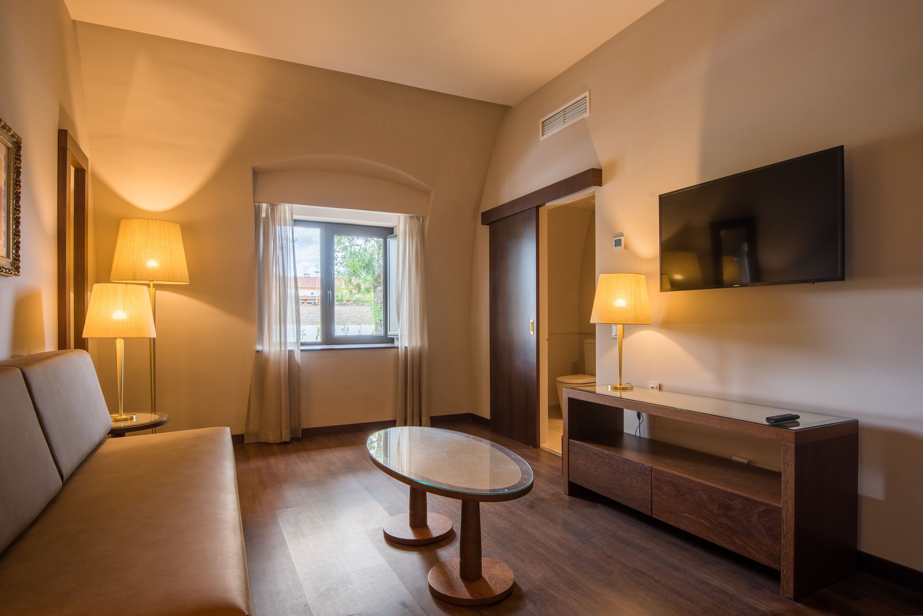 A hotel room with a sofa, a glass-top table, a TV mounted on the wall, and a desk with a lamp.