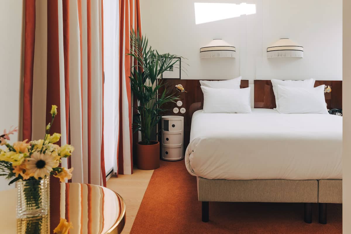 Ein Hotelzimmer mit einem großen Bett, einem Nachttisch, einer Zimmerpflanze und Vorhängen.