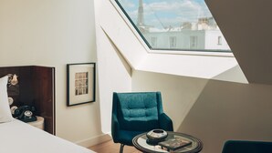 Habitación de hotel con cama, una silla, una mesita, un cuadro enmarcado y vistas de la Torre Eiffel a través de un tragaluz.