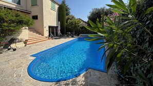 En swimmingpool med stenterasse, en trappe til en dør, og en bygning med beige facade og grønne skodder.