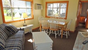 A cozy living room with a plaid sofa, a white coffee table, and a dining table with chairs. There are windows with blinds, an air conditioning unit, and a small table with a lamp.