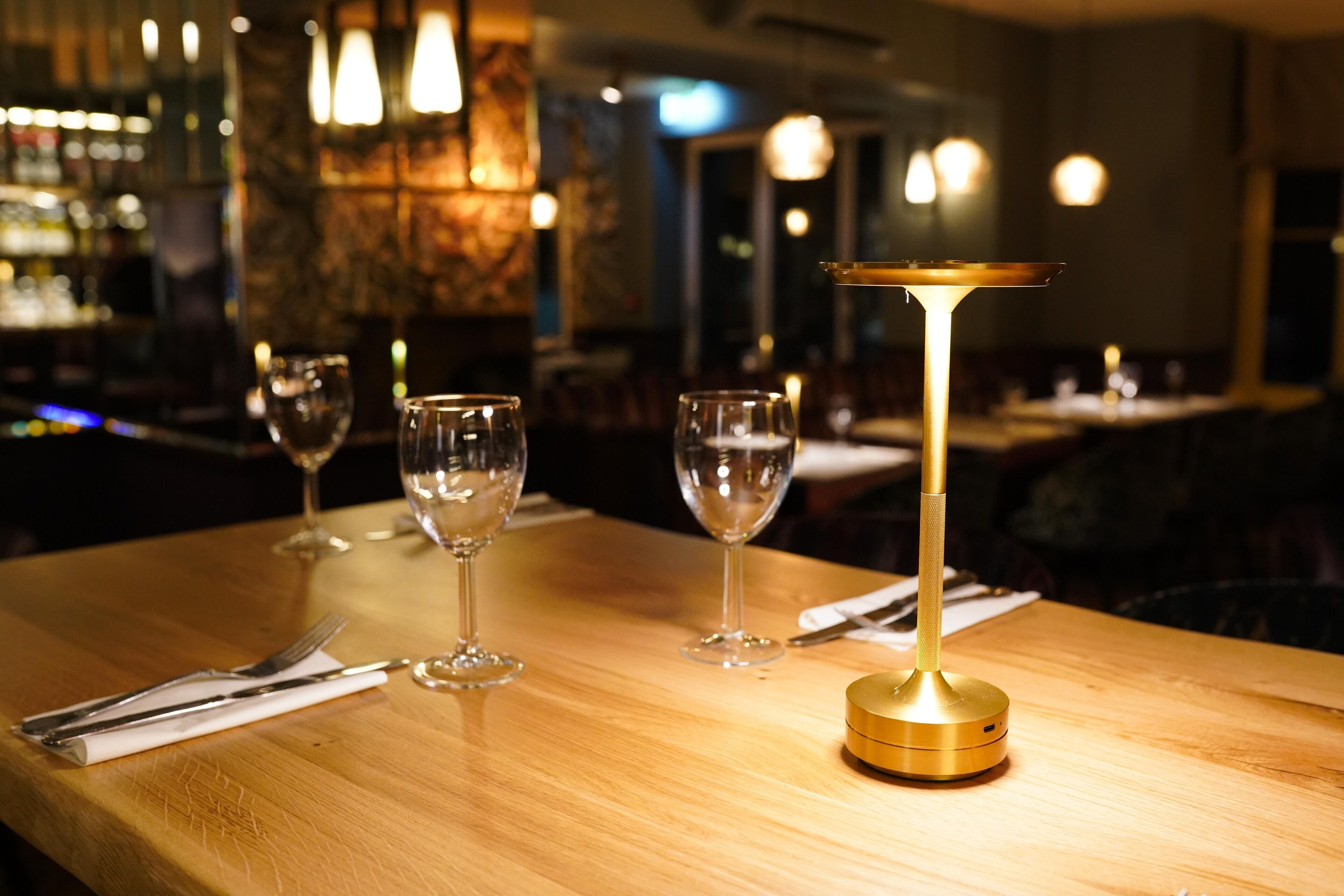 A bar setting with a wooden table, glasses, a gold-colored candle holder, and a dimly lit background with hanging lights.