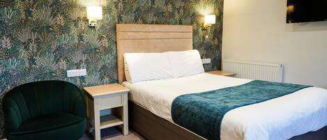 Habitación de hotel con cama, mesita de noche, sillón verde y televisor montado en la pared.
