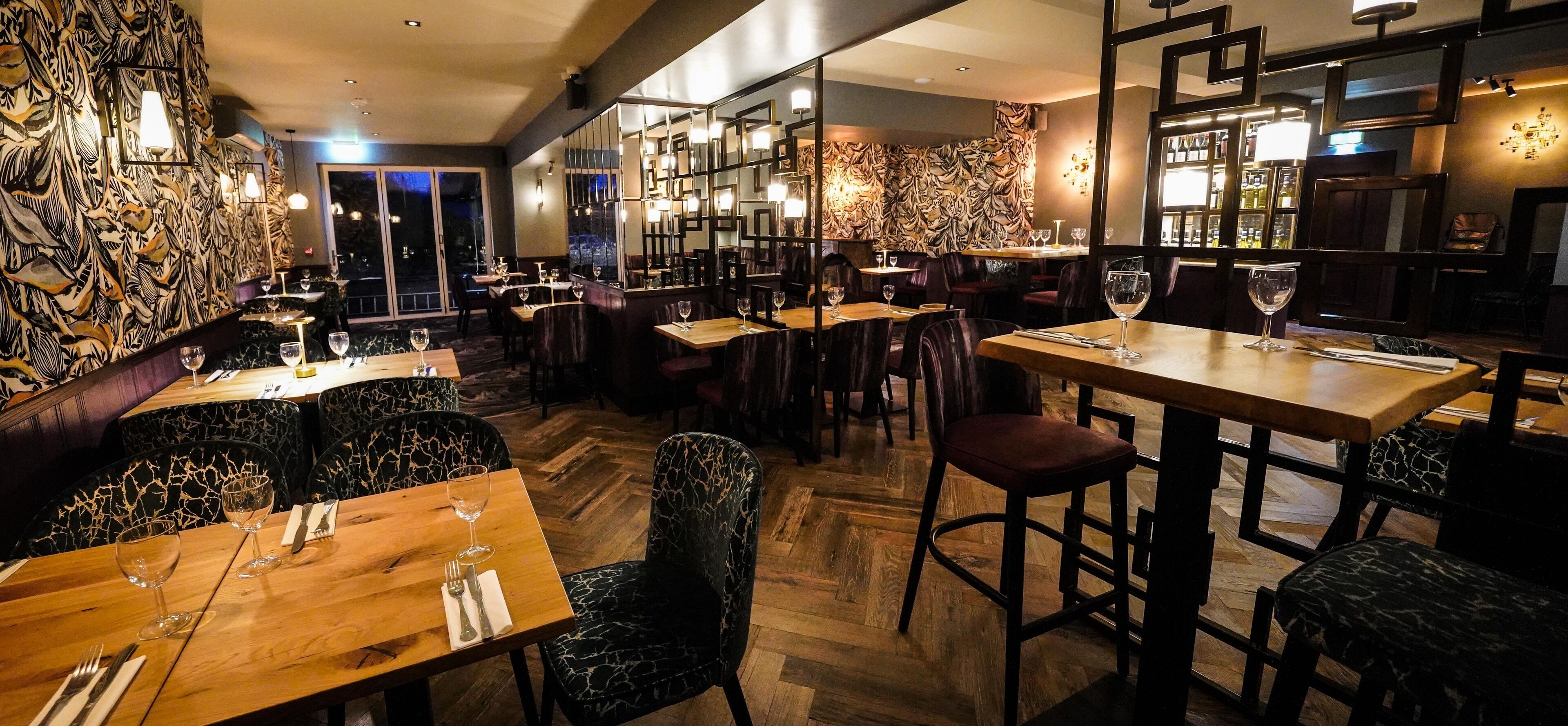 Interior de restaurant amb taules de fusta, cadires negres amb dibuixos i una zona de bar amb presentació de vi.