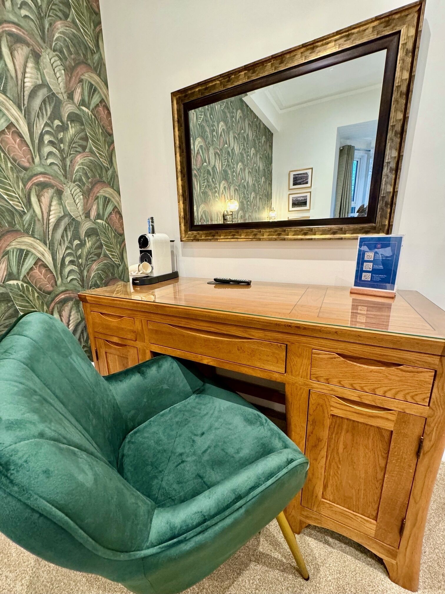 A wooden desk with a glass top, a green upholstered chair, and a large mirror above the desk.