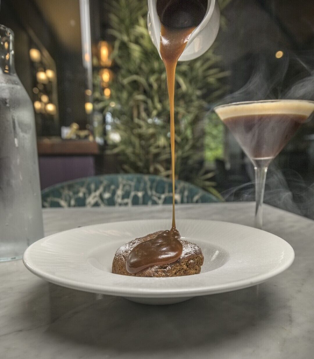 A dessert with a chocolate sauce being poured over it, a martini glass, and a bottle of water on a table.